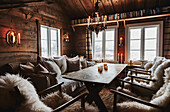 Gemütliches Wohnzimmer mit Kerzenlicht, bequemen Sitzgelegenheiten mit Kissen und Schaffell und Blick auf den Schnee durch die Fenster der Holzhütte in den Bergen von Sirdal, Norwegen