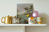 Krönungstassen und Milchkännchen mit Kunstwerken und goldener Teekanne auf einem Regal in einem Haus in Suffolk, England, Vereinigtes Königreich