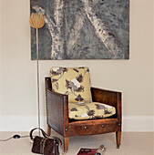 Vintage worn leather armchair with contemporary patterned cushions in a modern neutral living room with a standing reading lamp and a canvas oil painting