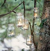 Recycled glass jam jars reused as candle holders hanging from tree branches in a wooded garden