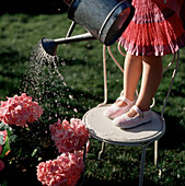 Junges Mädchen steht auf einem Metallgartenstuhl und gießt eine Pflanze
