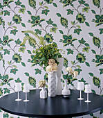 Grün gemusterte Tapete und schwarzer runder Esszimmertisch mit weißem Geschirr und einem Blumenarrangement
