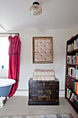 Framed fabric sample with folded quilts on oriental trunk and bookcase in bathroom of UK home