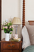 Cut flowers and lamp with books on wooden bedside table in Surrey barn conversion England UK