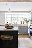 Wooden bar stool at marble topped kitchen island with sunlit window in Gloucestershire home UK