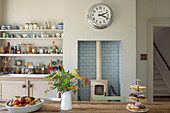 Cut flowers and cakes on table in kitchen with recessed wood burner and open shelving