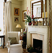 Armchair beside fireplace with pink orchids on mantlepiece and gilt mirror and pictures on the wall