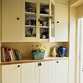Country style painted wooden kitchen units with open glass door and glazed pot planted with yellow Auriculars