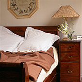 Close up of Mahogany bed with blanket and pillows and bedside cupboard with wicker lamp and flowers