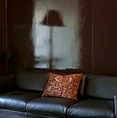 Floor lamp reflected in chocolate brown wall lacquer behind three seater leather divan with flocked cushion