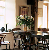 White flowers in glazed vase on oak dining table and chairs in country kitchen