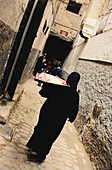 Frau trägt ein Tablett mit Lebensmitteln auf der Schulter in einer engen Gasse in der Medina von Fez, Marokko