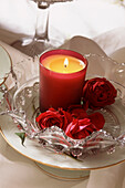 Lighted candle glowing on a ornate glass dish with red roses on a tabletop