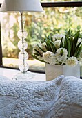 Close up of a white stitched bedspread with vase of white hyacinths and glass lamp stand by a window 