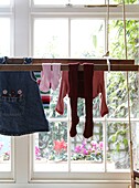 Kinderkleidung hängt auf einem Wäscheständer vor einem sonnendurchfluteten Fenster