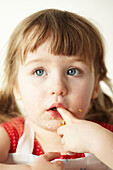 Three year old girl sits with finger in her mouth and blue eyes looking up