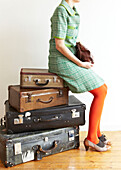 Woman in red tights and green dress sits on stack of vintage suitcases