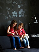 Two sisters sitting on sofa with floral patterned wallpaper in London townhouse, England, UK