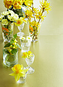 Yellow Daffodils, Primulas, Roses, and chocolate eggs in various vases on a gold table top