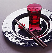 Karminroter Glasbecher und Essstäbchen auf glänzender versilberter Platte