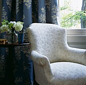 White armchair with pedestal side table and blue floral patterned curtains at window