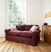 Mauve two-seater sofa with cushions on wooden floorboards at uncurtained window