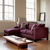 Mauve corner sofa with wooden coffee table in uncurtained room with sash windows