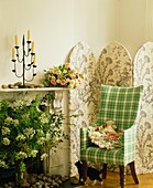 Checked green armchair with flower arrangement and folding screen at marble fireplace