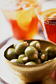 Rundes Tablett mit einer Schale grüner, mit Mandeln gefüllter Oliven und zwei Gläsern mit rosa Gin im Hintergrund