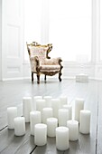 18 unlit church candles on painted floorboards in a minimally furnished all white room with bay window and antique armchair