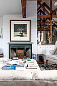 Framed artwork above fireplace with books on ottoman footstool in Norfolk coastguards cottage  England  UK