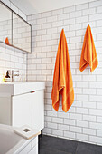Orange towels hang in white tiled London bathroom with mirrored cabinet above sink  UK