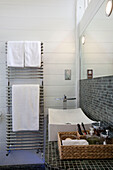 Zeitgenössisches Waschbecken und wandmontierter Heizkörper mit einem Korb mit Toilettenartikeln in einer grau gefliesten Nasszelle in einer umgebauten Wassermühle