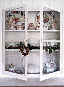 Glasvitrine mit offenen Türen, die eine geflieste Arbeitsfläche und eine mit altem Geschirr gefüllte und mit einer Stechpalmengirlande und einem Kranz geschmückte Wand zeigen