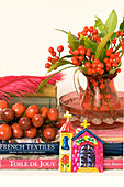 Still life with textile books feather cranberry glass jug filled with red berries and green leaves beside a pressed tin chapel decoration