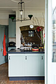 Spotted apron hangs on back of kitchen door with exposed brick recessed hob and pastel blue cupboards