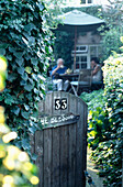 Open gate leading to cottage garden with people drinking tea at table