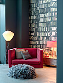 Pink armchair beside a wall decorated in bookcase wallpaper with grey floor cushion and floor lamp