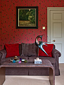 Vogelstatue auf Couchtisch im Wohnzimmer mit rot gemusterter Tapete und Kissen auf dem Sofa in einem Haus im ländlichen Suffolk, England, Vereinigtes Königreich