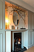 Zweigarrangement und ovaler Spiegel mit brennenden Kerzen am Kamin in einem Haus in Walberton, West Sussex, England, UK