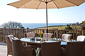 Wicker dining chairs at table with parasol on terrace, Cornwall, UK