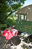Klappstühle und Tisch mit rot gepunkteter Plastiktischdecke auf schattiger Wohnwagen-Terrasse