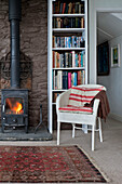 Bookshelf and lit burning stove with chair in beach house Cornwall England UK