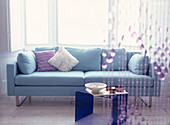 Pale blue sofa with cushions against a large bay window with purple beaded curtain hanging in the foreground