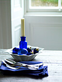 Candle in blue glass vase with cutlery and napkins on table