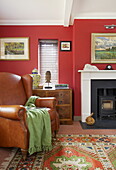 Brown leather armchair at fireplace in red living room of contemporary farmhouse, Nuthurst, West Sussex, England, UK