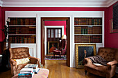 Pair of upholstered chairs in library with hardbacked books and gilt framed oil painting in Kent home England UK