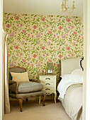 Upholstered armchair at bedside in Oxfordshire bedroom with floral patterned wallpaper, England, UK