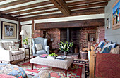 Armchairs and ottoman with exposed brick fireplace and beamed ceiling in living room of timber framed Kent cottage, England, UK