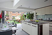 White and grey open plan kitchen and dining area with open doors to back garden in contemporary London home, England, UK